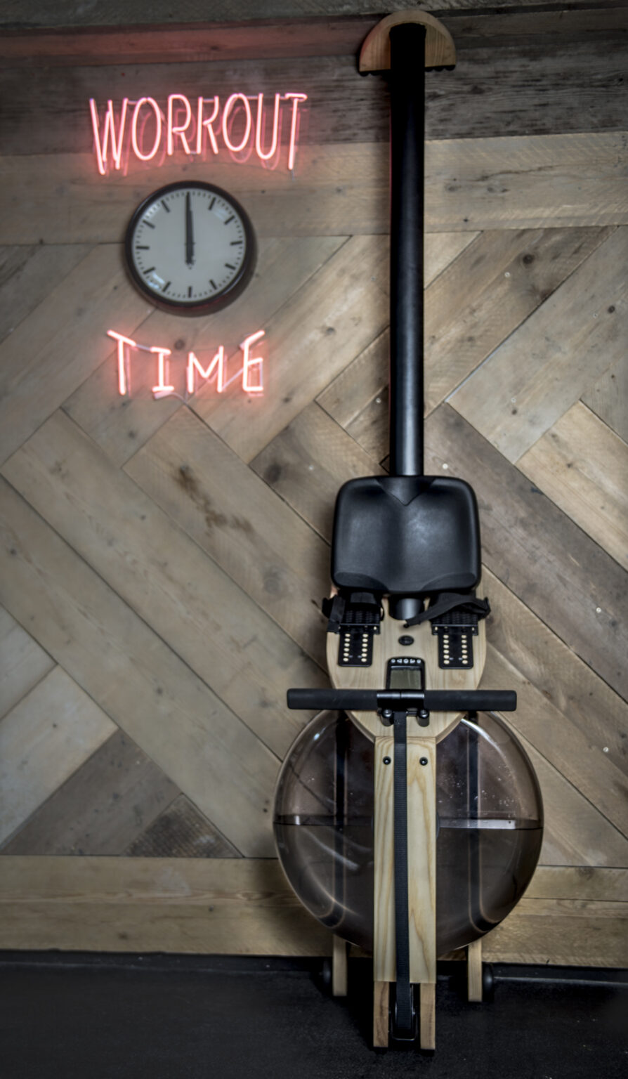 Hand-built wooden water rower in front of a chevron-pattern reclaimed timber wall, with a vintage clock and neon sign