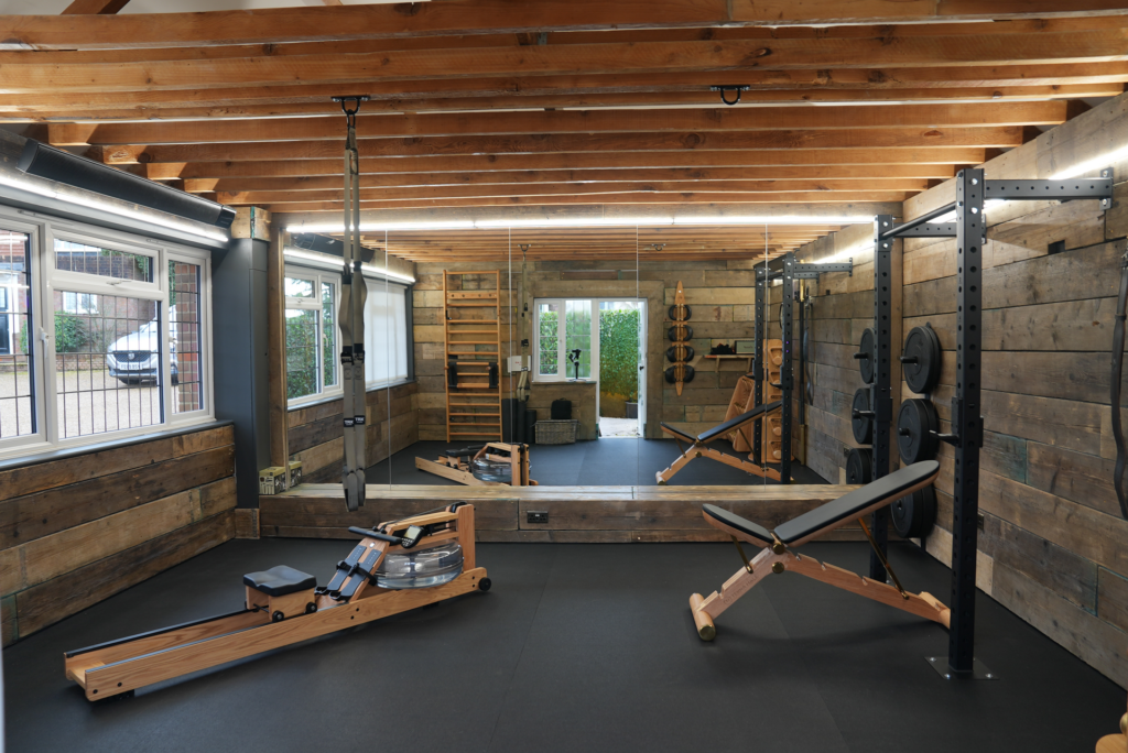 The same space rebuilt with reclaimed timber walls, exposed beam ceiling, wooden rowing machine and weight rack.