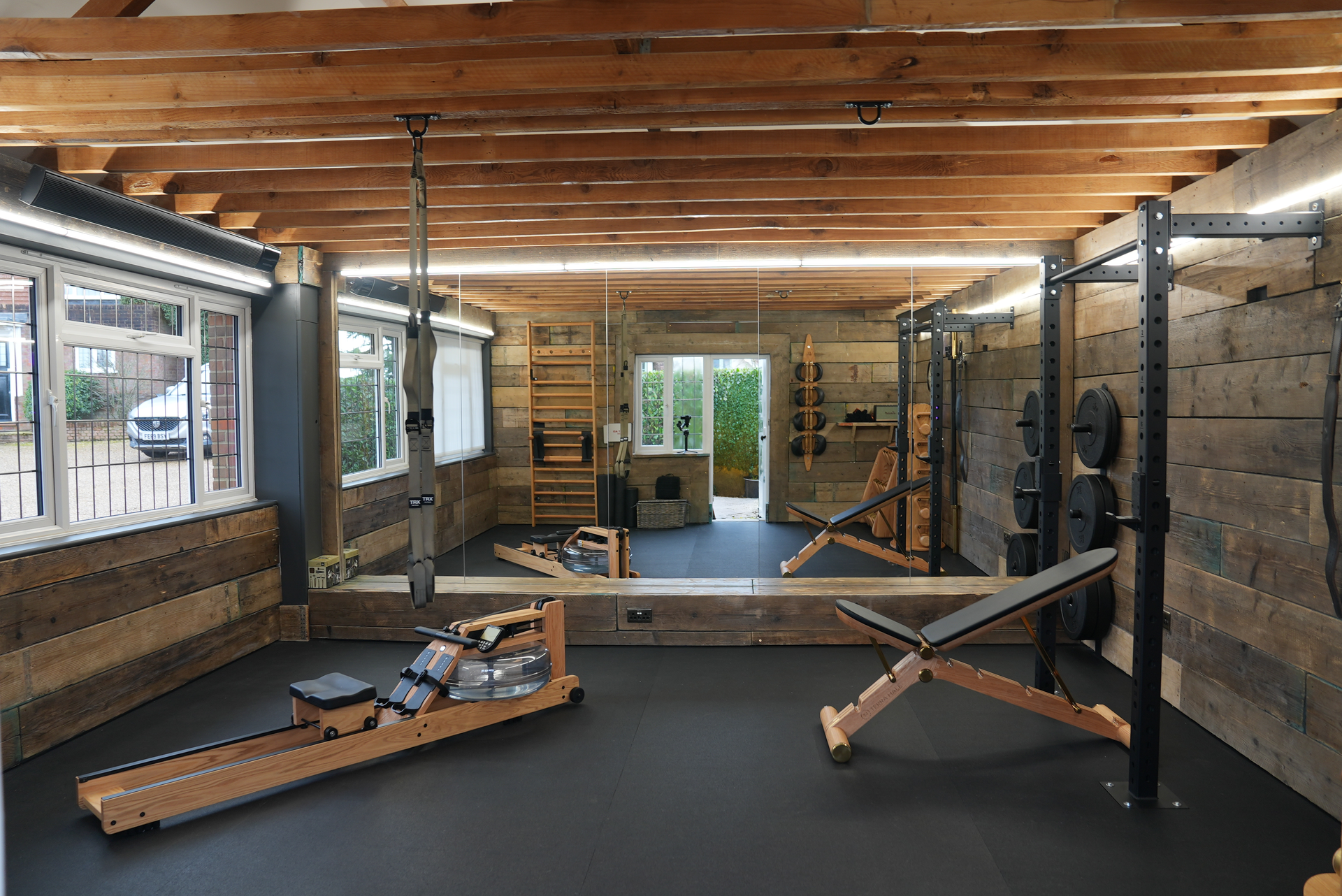 The same space rebuilt with reclaimed timber walls, exposed beam ceiling, wooden rowing machine and weight rack.